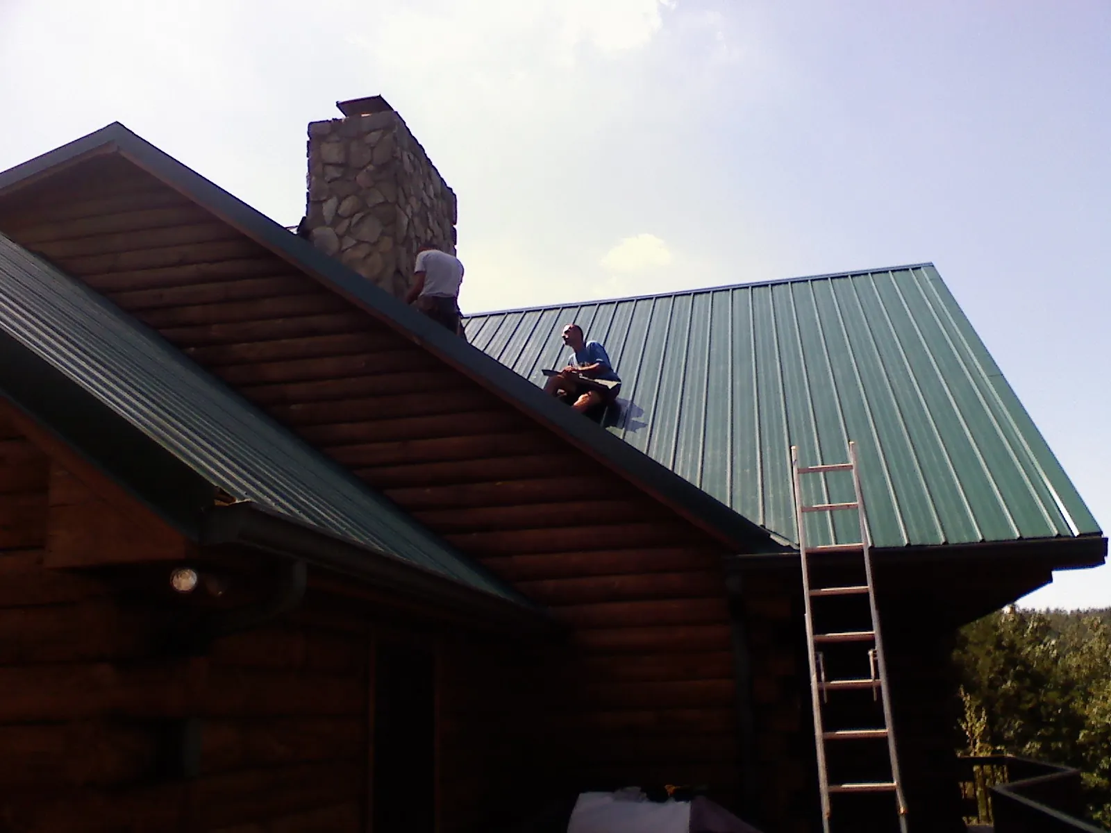 Roofers installing green metal panels beside a stone chimney on a log home in Arlington